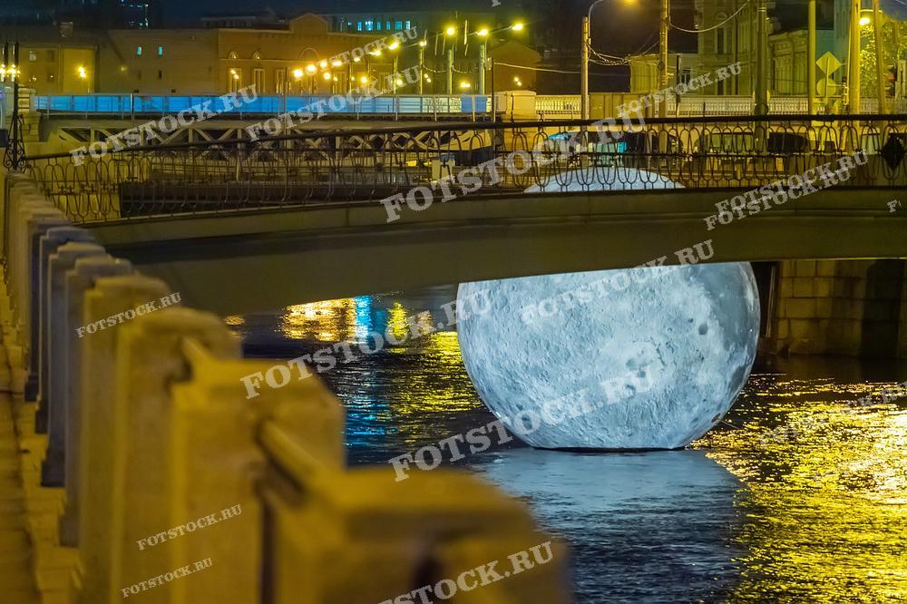 На фотографии изображена инсталляция - макет Луны, помещённый в Обводный канал. Такое необычное фото может быть использовано для украшения интерьеров, в культурно-исторических публикациях о городе или в рекламе ночных городских туров.